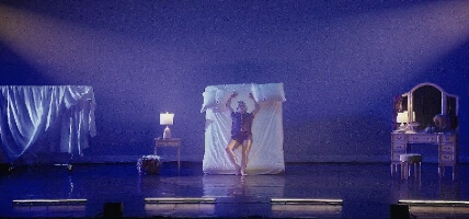 On a large stage, there is a vanity with a lamp and 2 mannequin heads with wigs. In the center of the stage, a Latina woman in her 30s is leaning and posing with her arms open on a bed that has been tilted up vertically. She is dressed in silk pajamas and has a head wrap.