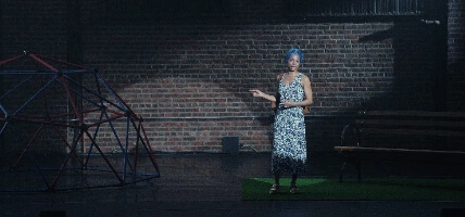 A Black woman wearing a head wrap and a flowing blue dress stands on stage with a park bench and a geometric climbing dome. She is looking straight forward with concern.
