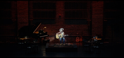 A white woman in her 50s is sitting in a chair on stage that is set like a coffee shop with tables and chairs. She is playing a guitar and is singing into a microphone.